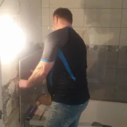 Glazurnik układa szarą płytkę na ścianie łazienki z widocznymi śladami po poprzednich płytkach, podświetlona żarówką, w tle wanna i fragment ułożonych płytek z dekoracyjnym pasem.