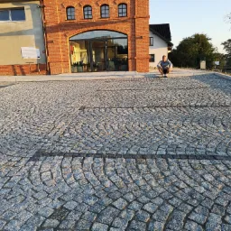 Świeżo ułożona, szara kostka granitowa w kształcie wachlarza przed budynkiem z czerwonej cegły. Mężczyzna w dresie siedzi na krawężniku. Widoczny fragment maszyny budowlanej.