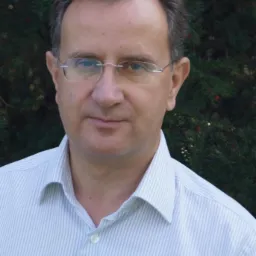 Portret mężczyzny w okularach i koszuli w paski na tle ciemnozielonych iglaków.