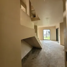 Surowe wnętrze domu w trakcie prac wykończeniowych, ściany i sufit pokryte świeżym tynkiem, widoczne schody w stanie surowym, na podłodze resztki budowlane, przewody zwisające z sufitu.