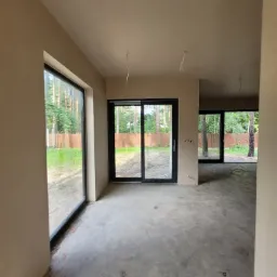 Wnętrze domu po nałożeniu jasnego tynku, widoczne duże okna przesuwne z czarnymi ramami i surowy beton na podłodze, przygotowane do dalszych prac wykończeniowych.