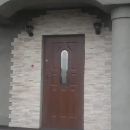 Wejście do domu z brązowymi drzwiami i elewacją wykończoną jasnym kamieniem dekoracyjnym, oświetlone dwoma lampami ściennymi.