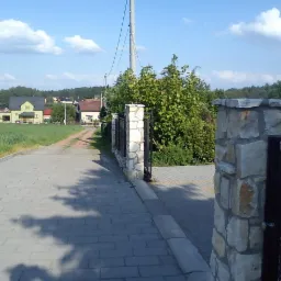 Kamienny słupek ogrodzeniowy z metalową bramą w tle krajobrazu wiejskiego z domami i polami.