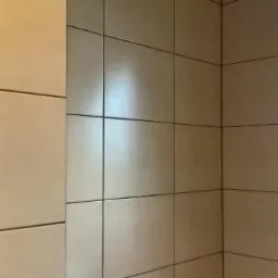 Wykończona ściana w łazience z beżowymi, kwadratowymi płytkami ceramicznymi i ciemnymi fugami, widoczny narożnik i odbicie światła na powierzchni płytek.