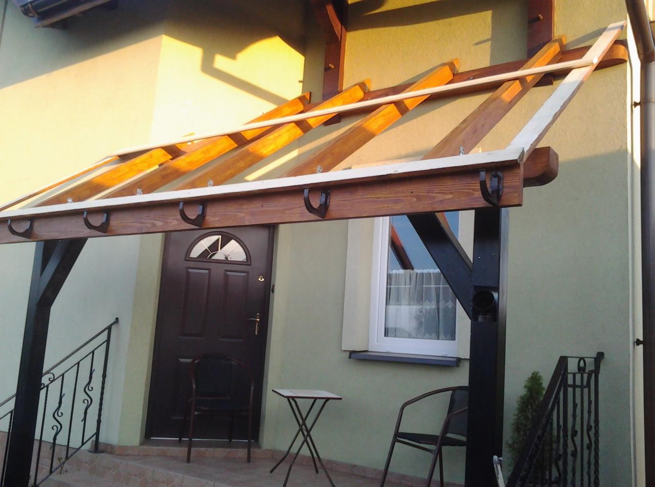 Zadaszenie nad wejściem do domu z drewnianą konstrukcją, brązowe drzwi wejściowe i okno z firanką w tle, zewnętrzne oświetlenie.