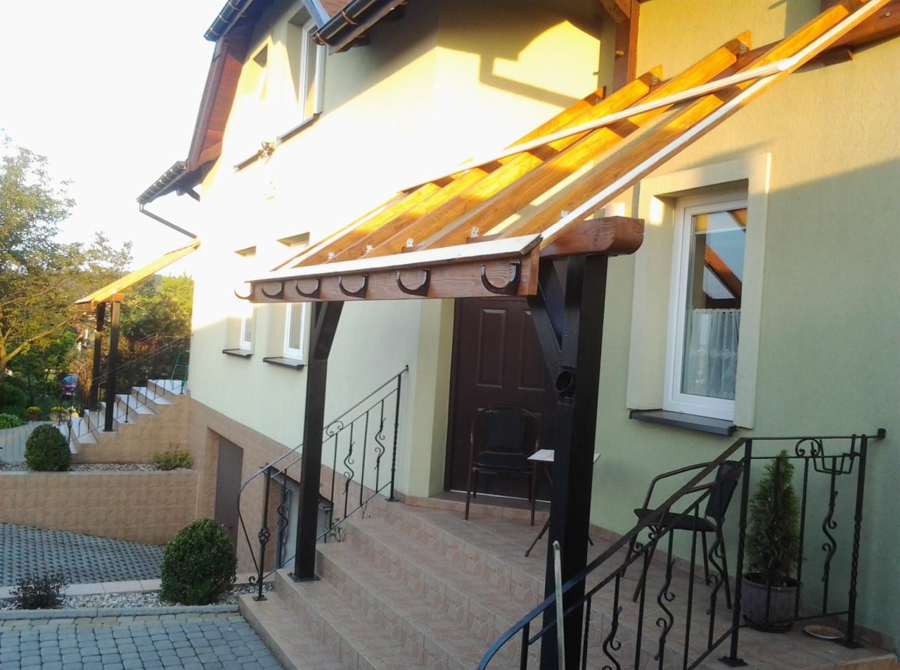 Zadaszenie nad wejściem do domu z drewnianą konstrukcją i ozdobnymi rynnami, pomalowane na jasny kolor elewacja i czarne elementy metalowe, widoczne schody i balustrady.