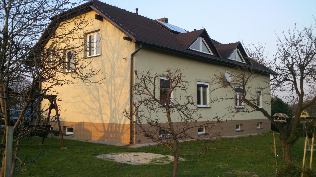 Dwukondygnacyjny dom z beżową elewacją, brązowym dachem i panelami słonecznymi, widoczny wczesną wiosną z drzewami bez liści i huśtawką na trawniku.