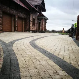 Ułożona kostka brukowa w dwóch odcieniach szarości tworząca wzór przed domem z cegły, pracownik w odblaskowej kamizelce docina kostkę przy ogrodzeniu.