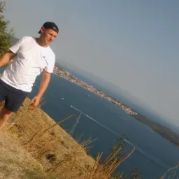 Mężczyzna w czapce i białej koszulce stoi na wzgórzu z widokiem na morze i miasto w tle, w pobliżu słupów elektrycznych.