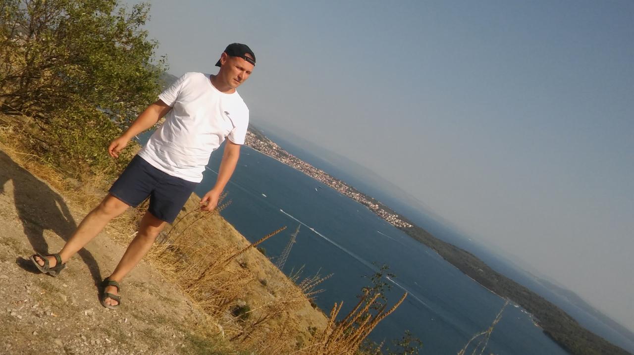 Mężczyzna w czapce i białej koszulce stoi na wzgórzu z widokiem na morze i miasto w tle, w pobliżu słupów elektrycznych.