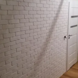 Ściana wykończona białymi płytkami imitującymi cegłę, z widocznym fragmentem niezakończonej pracy przy suficie, obok białe drzwi z poziomymi przeszkleniami i drewniana podłoga.