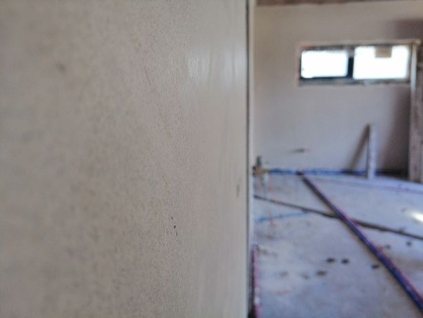 Świeżo nałożony, gładki tynk na ścianie w surowym wnętrzu z widocznymi instalacjami hydraulicznymi i oknem w tle.