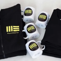 Czarna bluza z zamkiem błyskawicznym, stos czarnych koszulek i cztery białe kubki z nadrukiem logo #muzoedukacja, ułożone na jasnym tle.