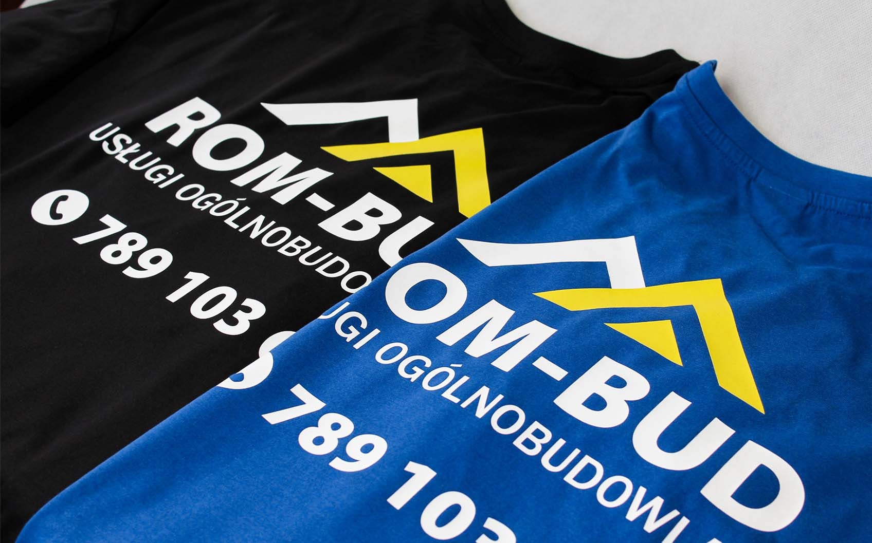 Dwa t-shirty: czarny i niebieski, z nadrukowanym logo firmy budowlanej 'ROM-BUD' wraz z numerem telefonu, leżące na jasnym tle.