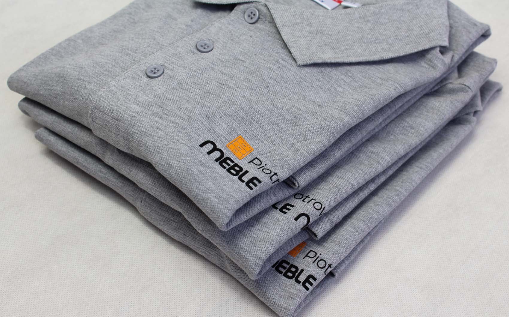 Stos szarych koszulek polo z nadrukiem logo firmy meblarskiej, widoczne trzy guziki i kołnierzyk, leżące na białym tle.