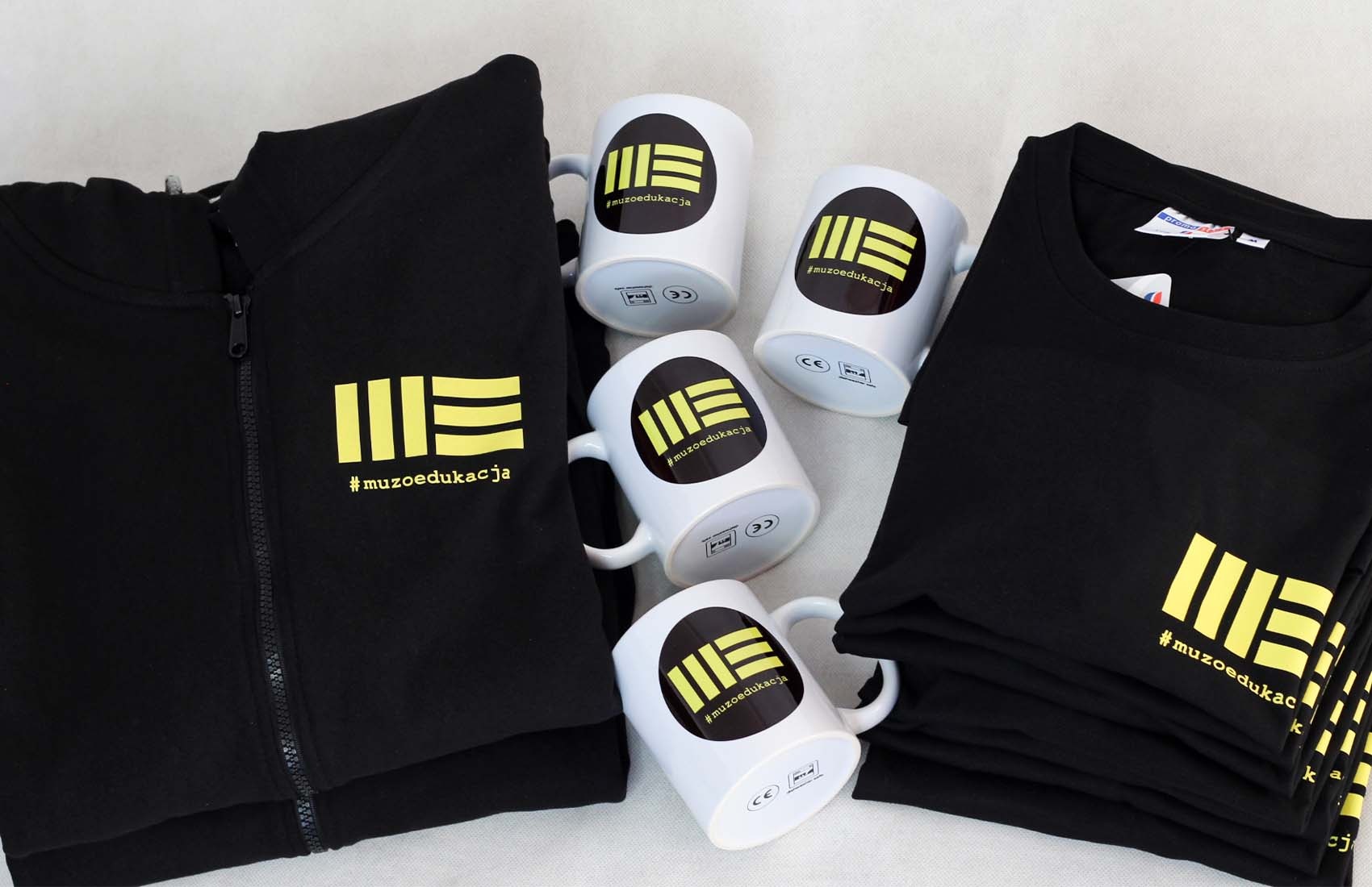 Czarna bluza z zamkiem błyskawicznym, stos czarnych koszulek i cztery białe kubki z nadrukiem logo #muzoedukacja, ułożone na jasnym tle.