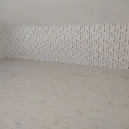 Poddasze po remoncie: świeżo położone panele podłogowe w odcieniach bieli i ściana wykończona tapetą w drobny wzór strzałek, widok na skos pomieszczenia.