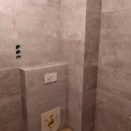 Szara łazienka w trakcie wykańczania, widoczne płytki imitujące kamień, podtynkowa instalacja WC z żółtymi elementami i otwory na baterie.