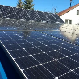 Panele fotowoltaiczne zamontowane na dachu budynku w Olsztynie, widoczne z bliskiej perspektywy pod kątem, z budynkiem w tle i fragmentem anteny.