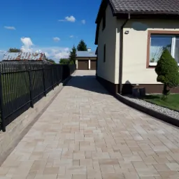 Ułożona beżowa kostka brukowa na podjeździe do domu jednorodzinnego z czarnym ogrodzeniem, widok w kierunku garażu i błękitnego nieba.