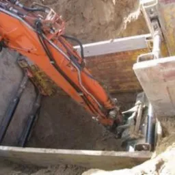 Pomarańczowa koparka zagłębia ramię w wykop zabezpieczony szalunkami, widoczne elementy hydrauliczne i ziemia wewnątrz wykopu.