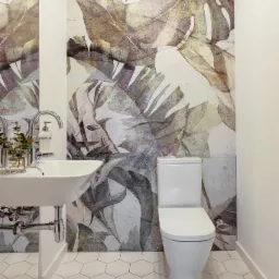 Nowoczesna toaleta z umywalką na tle ściany z motywem liści monstery w odcieniach szarości i beżu, biała armatura i heksagonalne płytki podłogowe.