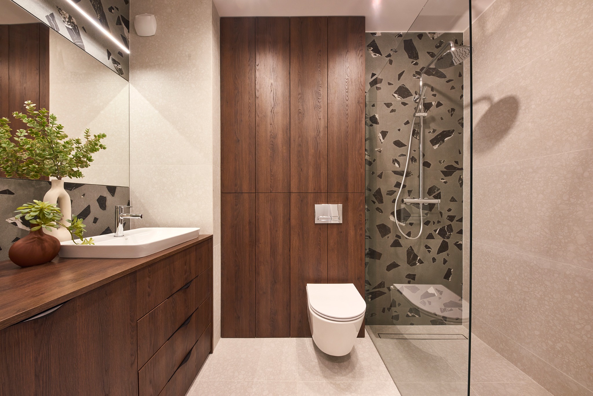 Nowoczesna łazienka z drewnianą szafką pod umywalką, wiszącą toaletą i kabiną prysznicową z płytkami w geometryczne wzory, oświetlenie LED w lustrze.