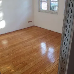 Błyszcząca, świeżo polakierowana podłoga z desek w pustym pokoju, widoczne gniazdka elektryczne na ścianie i fragment metalowej konstrukcji po prawej stronie.