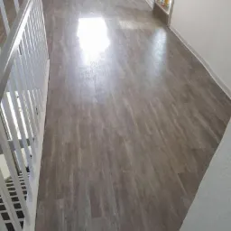 Nowo ułożona podłoga z paneli imitujących drewno, widoczna z perspektywy schodów z białą balustradą, z fragmentem białej ściany i grzejnika w tle.