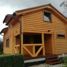 Gotowy, parterowy budynek z bali drewnianych, z zadaszonym wejściem i widoczną konstrukcją dachu oraz brązowymi rynnami, na tle pochmurnego nieba.