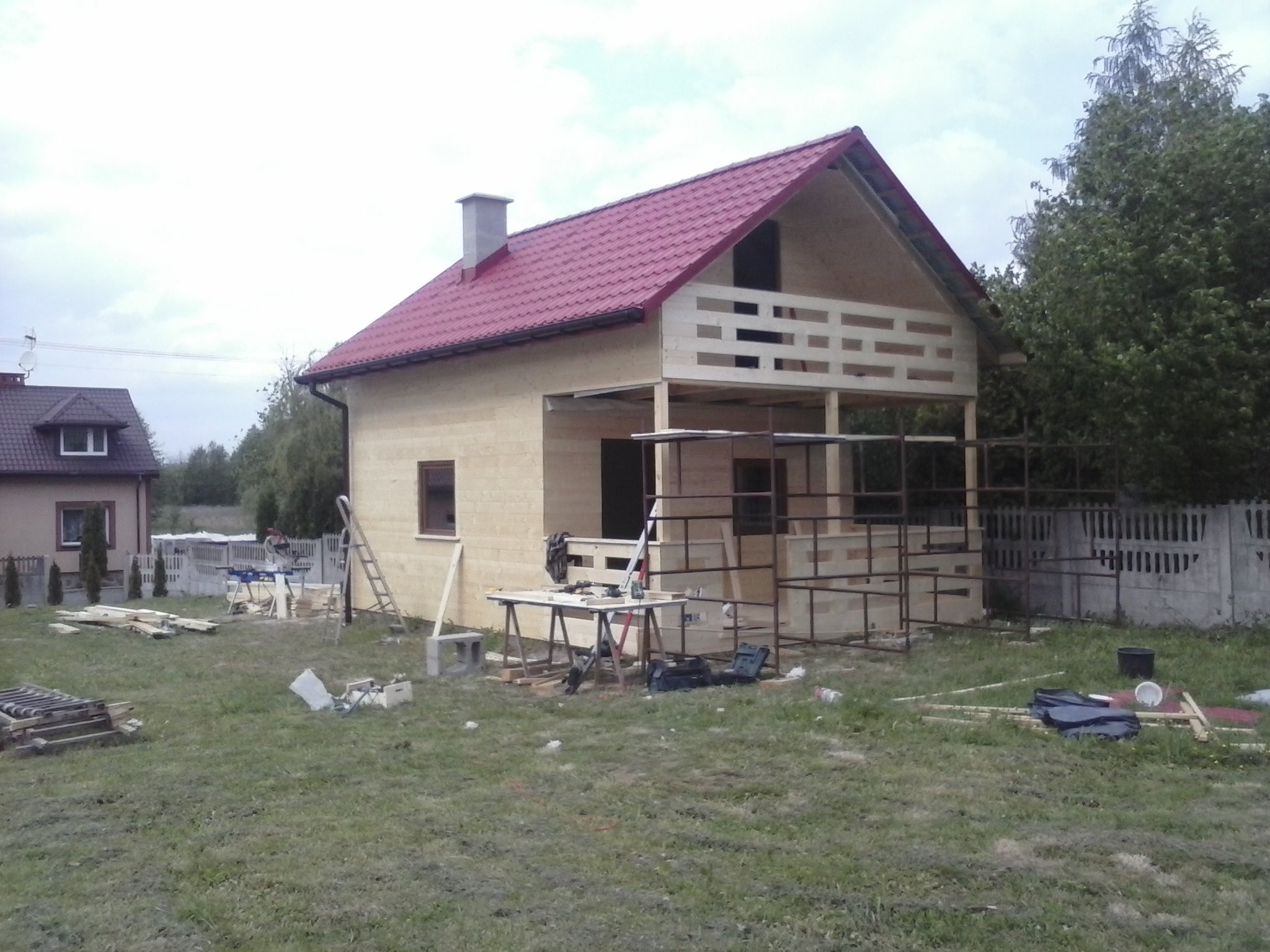 Wznoszony budynek z czerwonym dachem, widoczna konstrukcja drewniana balkonu, narzędzia rozłożone na trawie, obok drugi dom i betonowe ogrodzenie.