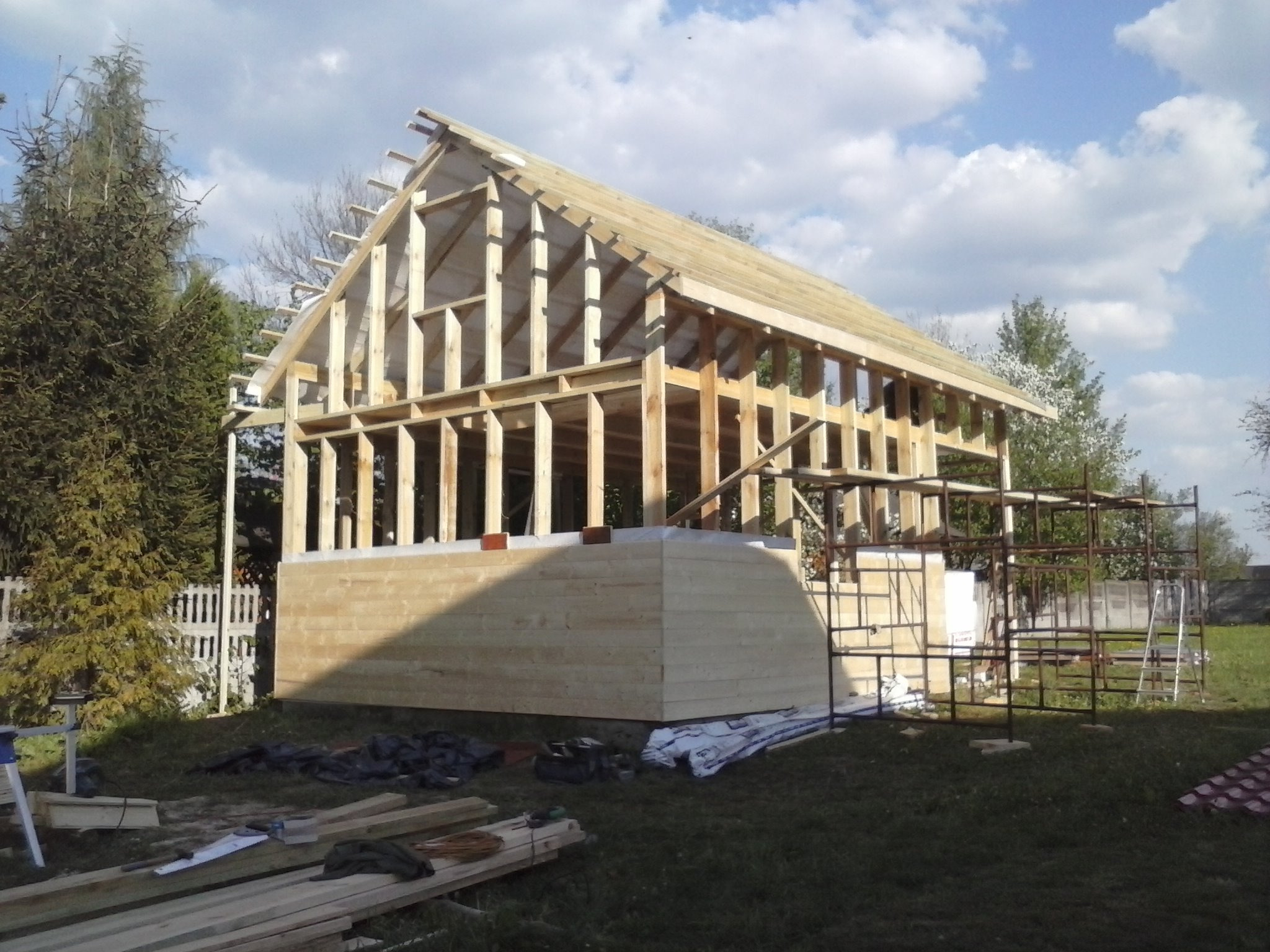 Budowa domu szkieletowego w trakcie realizacji: widoczna drewniana konstrukcja ścian i dachu, częściowo obłożona deskami, z elementami rusztowania i materiałów budowlanych na trawiastym terenie.