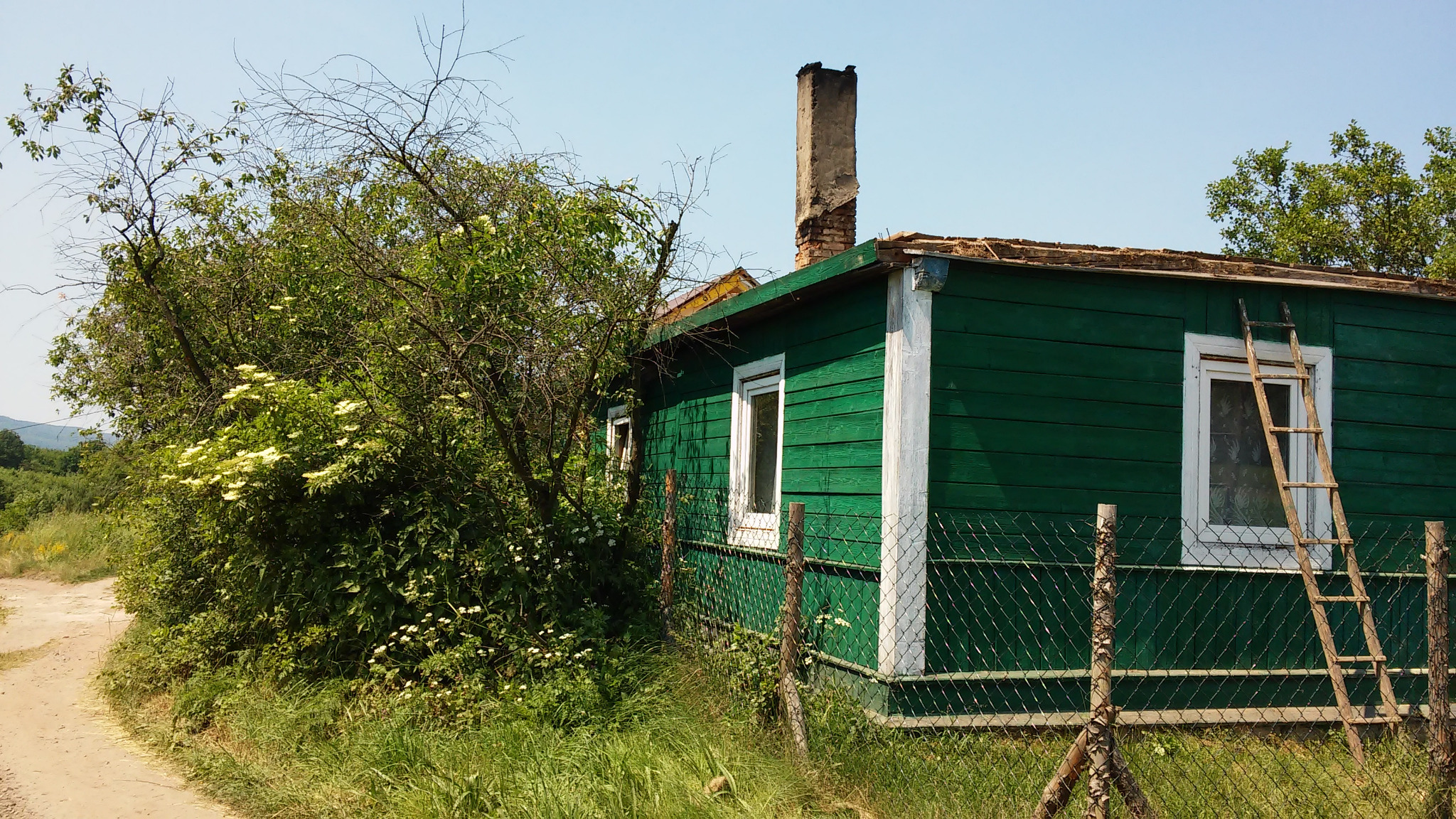 Zielony, drewniany budynek z drabina opartą o okno, widoczny z zewnątrz przez siatkę ogrodzeniową, z uszkodzonym kominem i częściowo zerwanym poszyciem dachu.