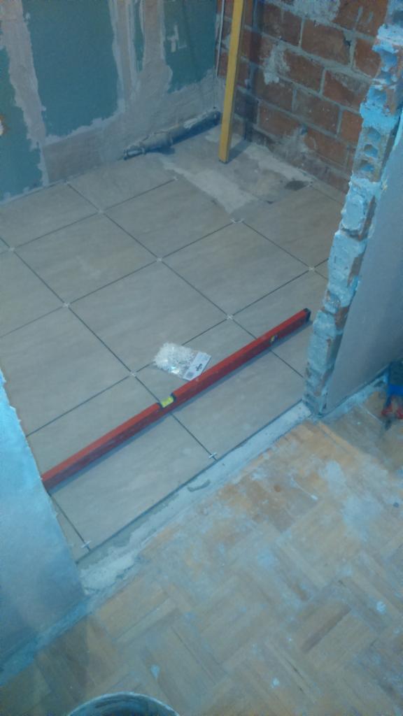 Układanie beżowych płytek podłogowych w łazience, z użyciem poziomicy i krzyżyków dystansowych; widoczny fragment surowej ściany z cegły i stara podłoga parkietowa.