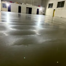 Wnętrze hali z nową, błyszczącą posadzką przemysłową w odcieniach szarości. Widoczne refleksy światła na gładkiej powierzchni i fragmenty ścian z oknami.