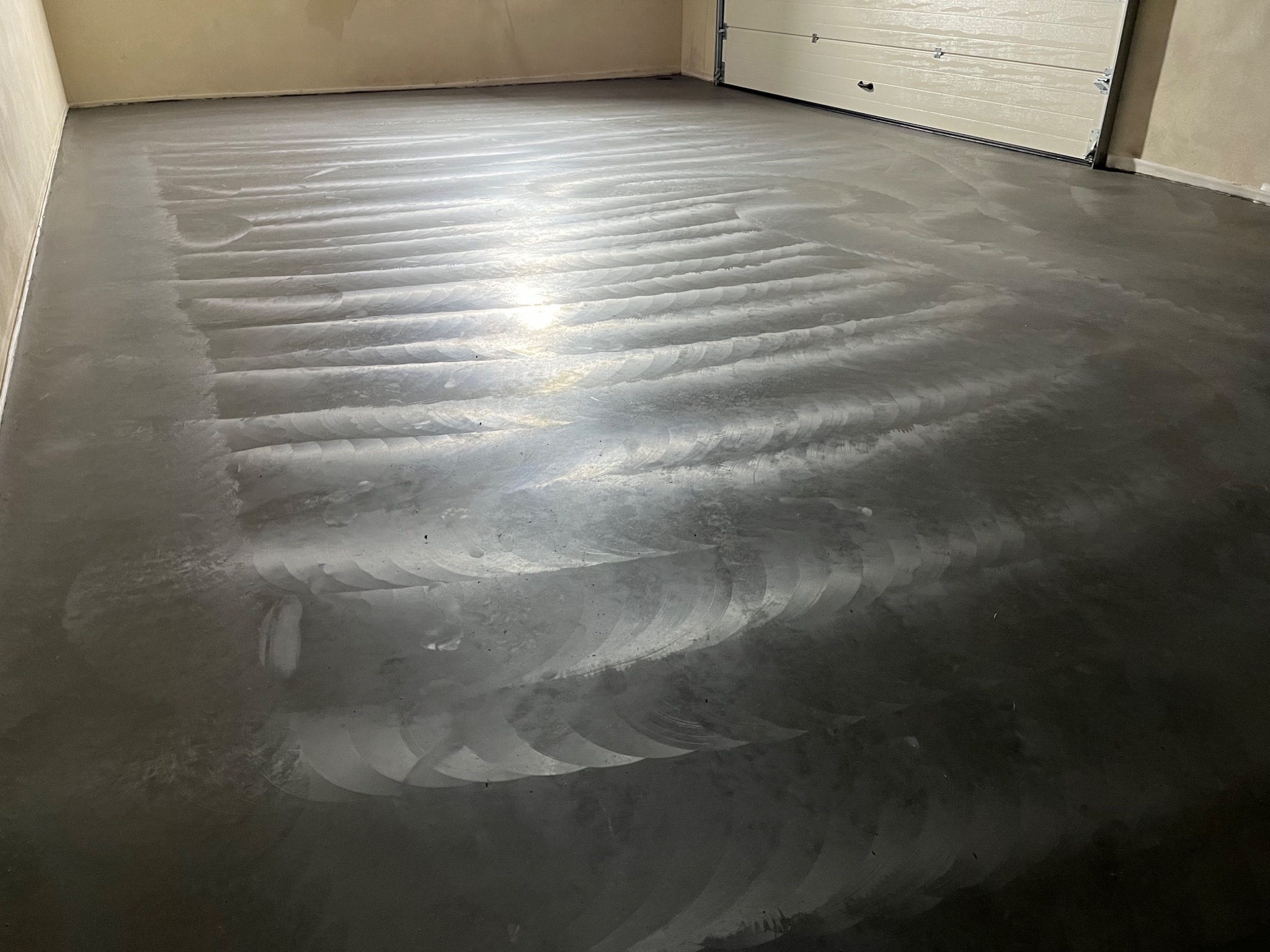 Świeżo wylana, szara posadzka w garażu z widocznymi śladami po zacieraniu, odbijająca światło, z białą bramą garażową w tle.