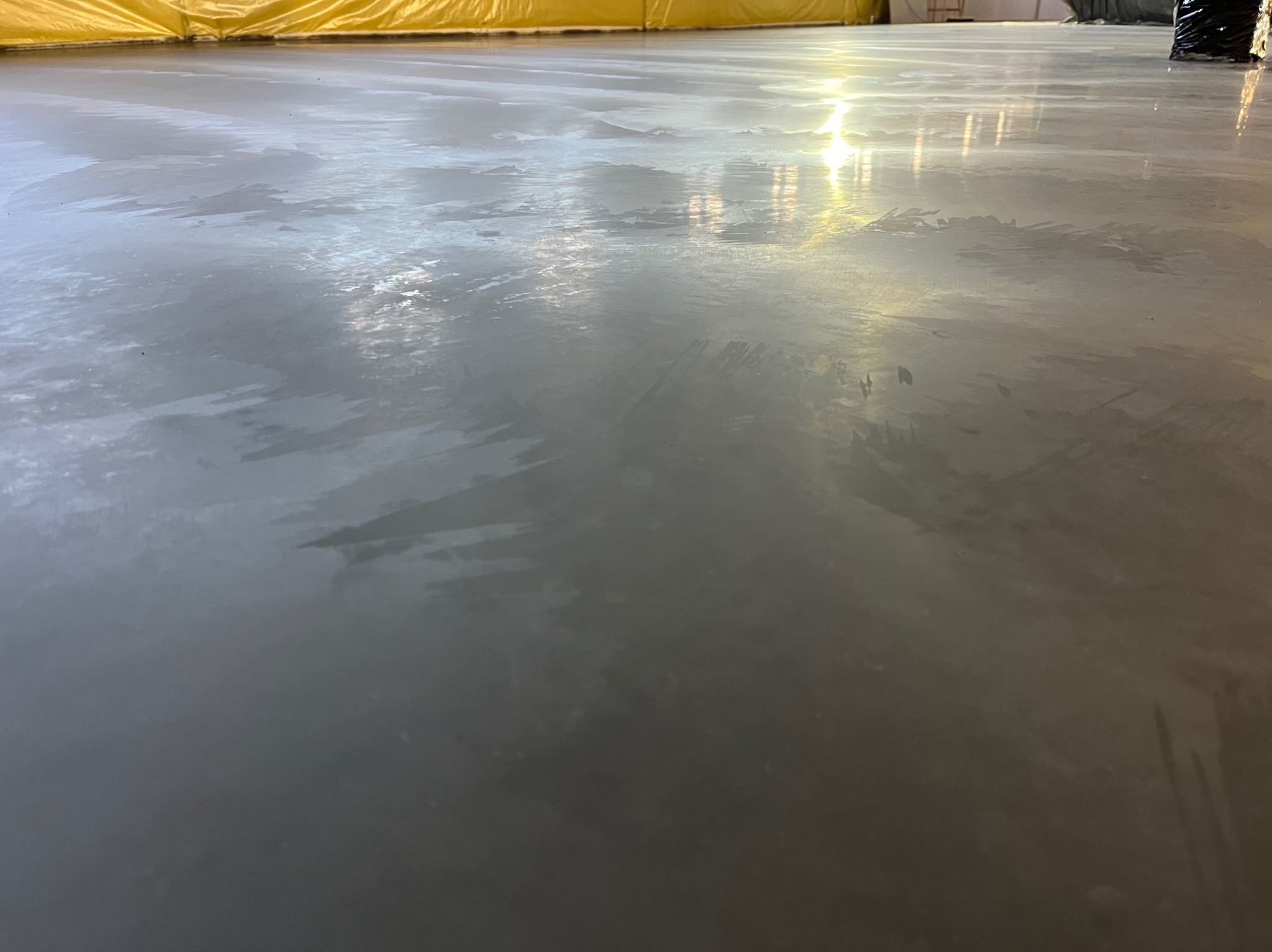 Świeżo wylana, mokra, szara posadzka z widocznymi smugami i refleksami światła, zabezpieczona żółtą folią ochronną przy ścianie.