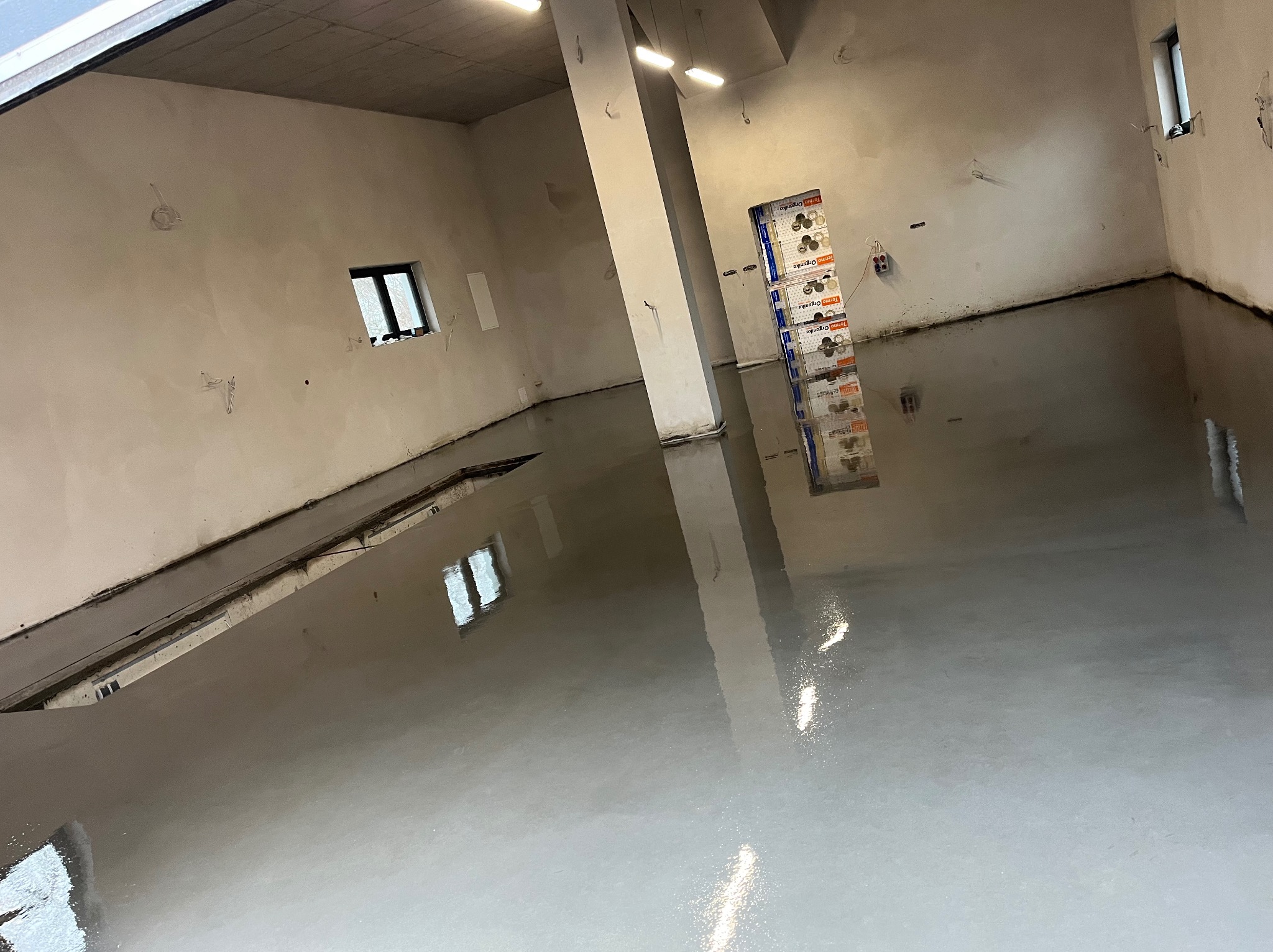 Świeżo wylana, samopoziomująca szara posadzka w surowym wnętrzu hali, z widocznymi refleksami światła i stosem materiałów budowlanych w tle.