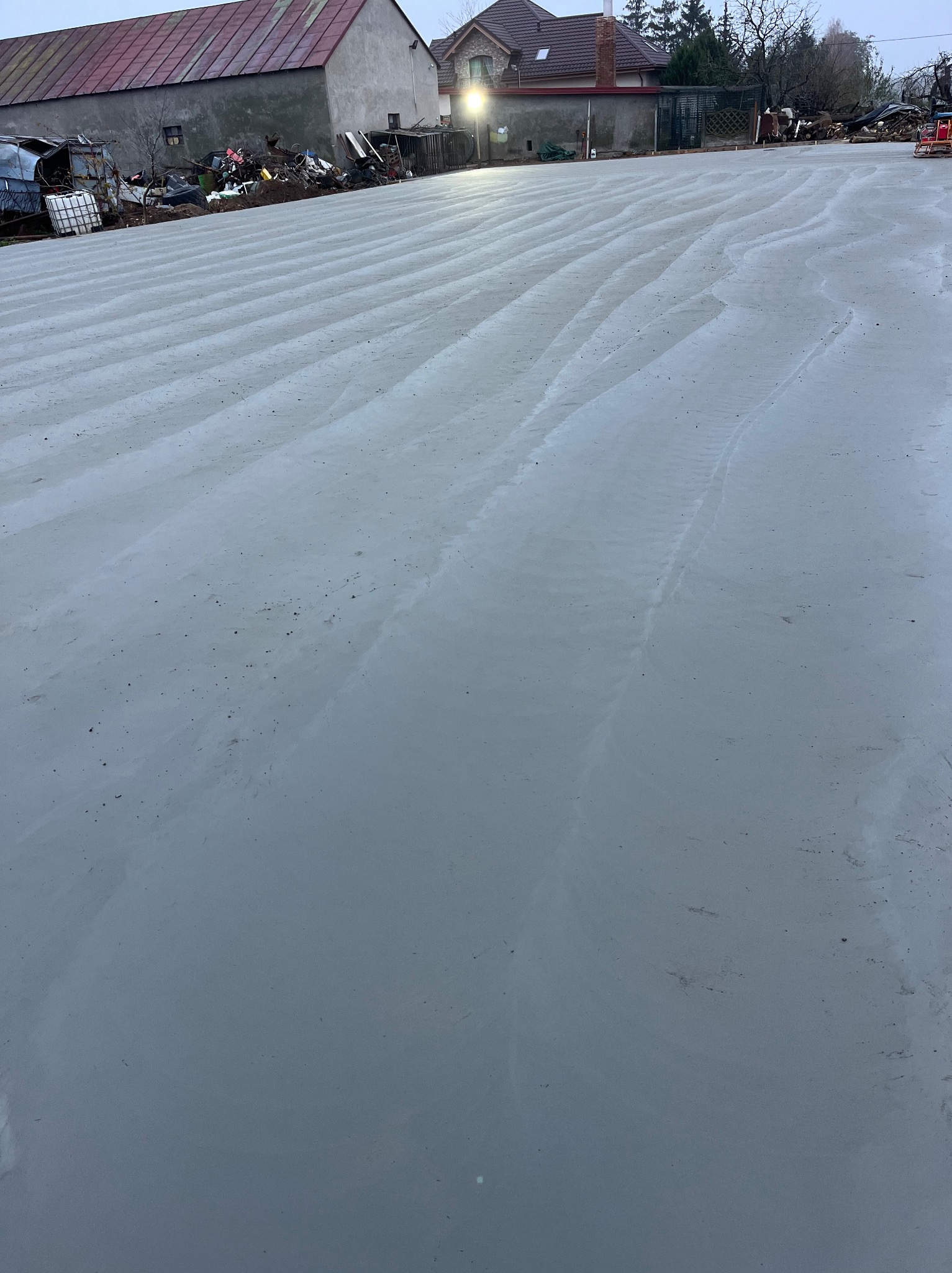 Świeżo wylana, gładka, szara betonowa posadzka z delikatnymi śladami po narzędziach, w tle budynki gospodarcze i gruzowisko.