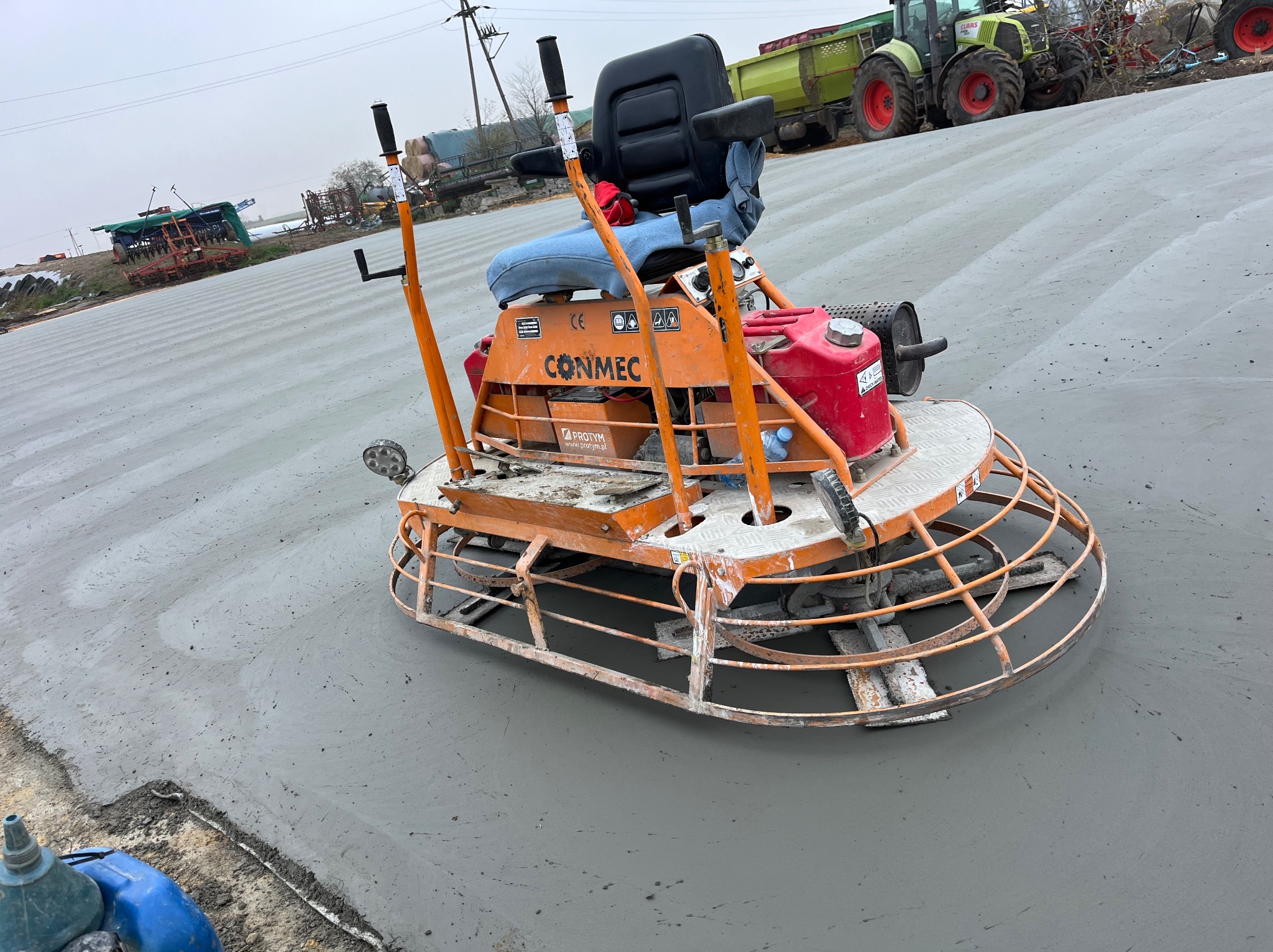 Zacieraczka mechaniczna Conmec na świeżo wylanej, szarej posadzce przemysłowej, w tle traktor i inne maszyny rolnicze na pochmurnym niebie.
