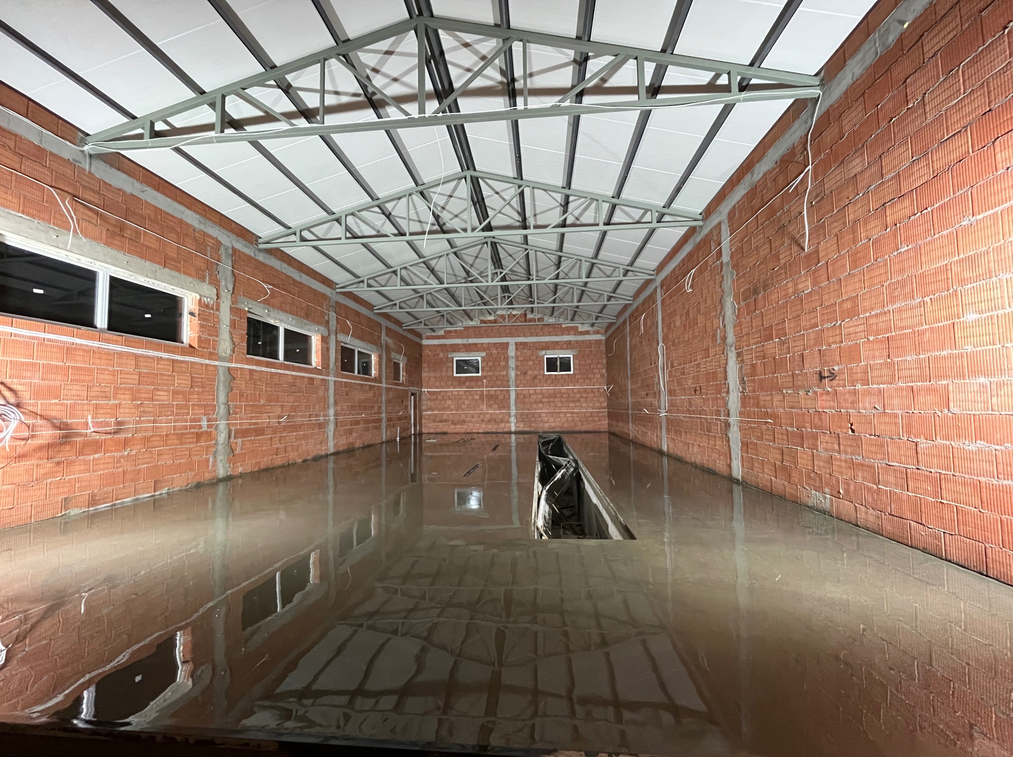 Świeżo wylana, mokra posadzka w surowym, ceglanym wnętrzu hali, z widocznymi refleksami światła i metalową konstrukcją dachu.