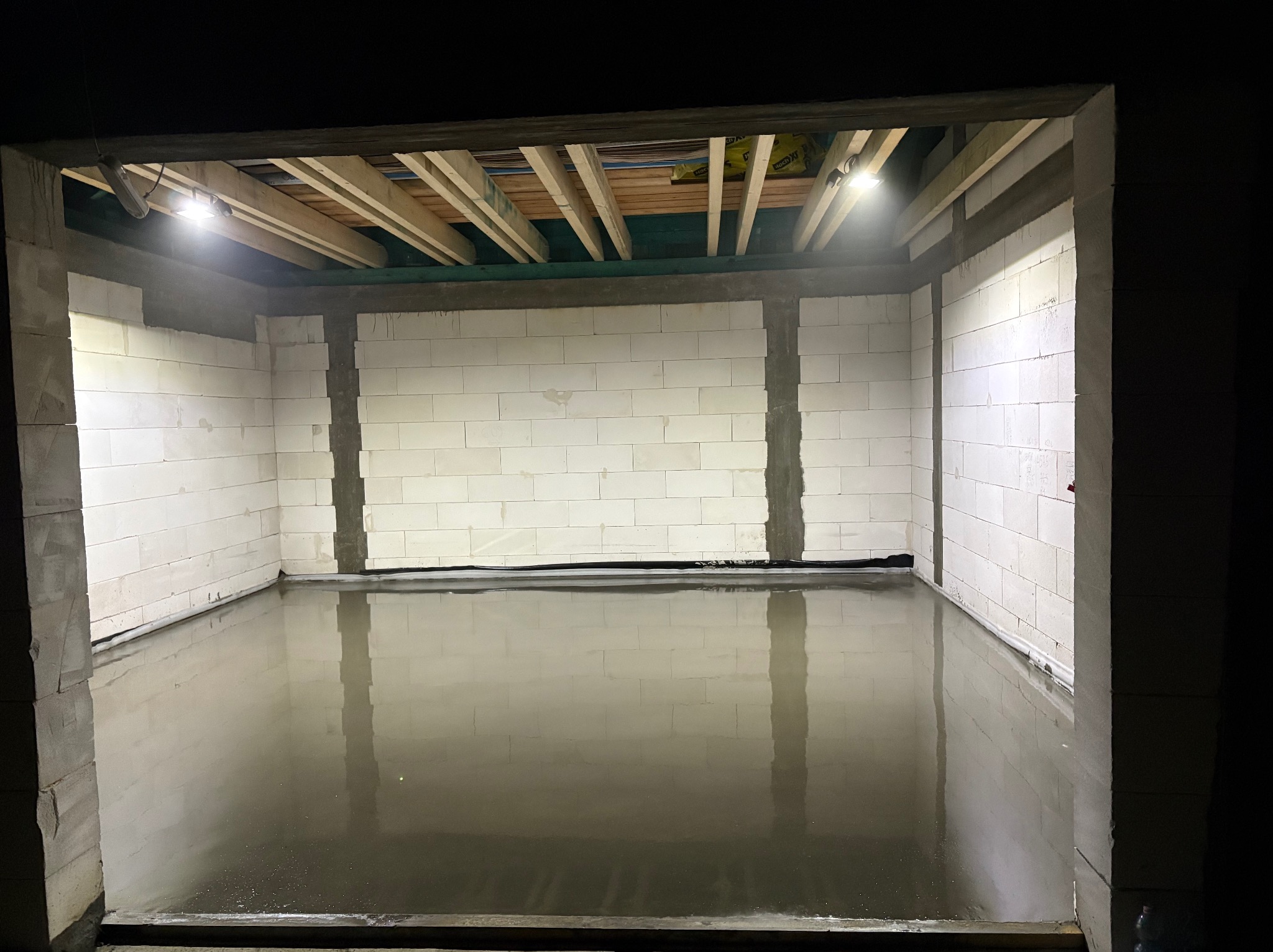 Świeżo wylana, mokra posadzka w pomieszczeniu z białymi ścianami z bloczków w Szczutowie, oświetlona lampami budowlanymi, z widoczną konstrukcją drewnianą stropu.