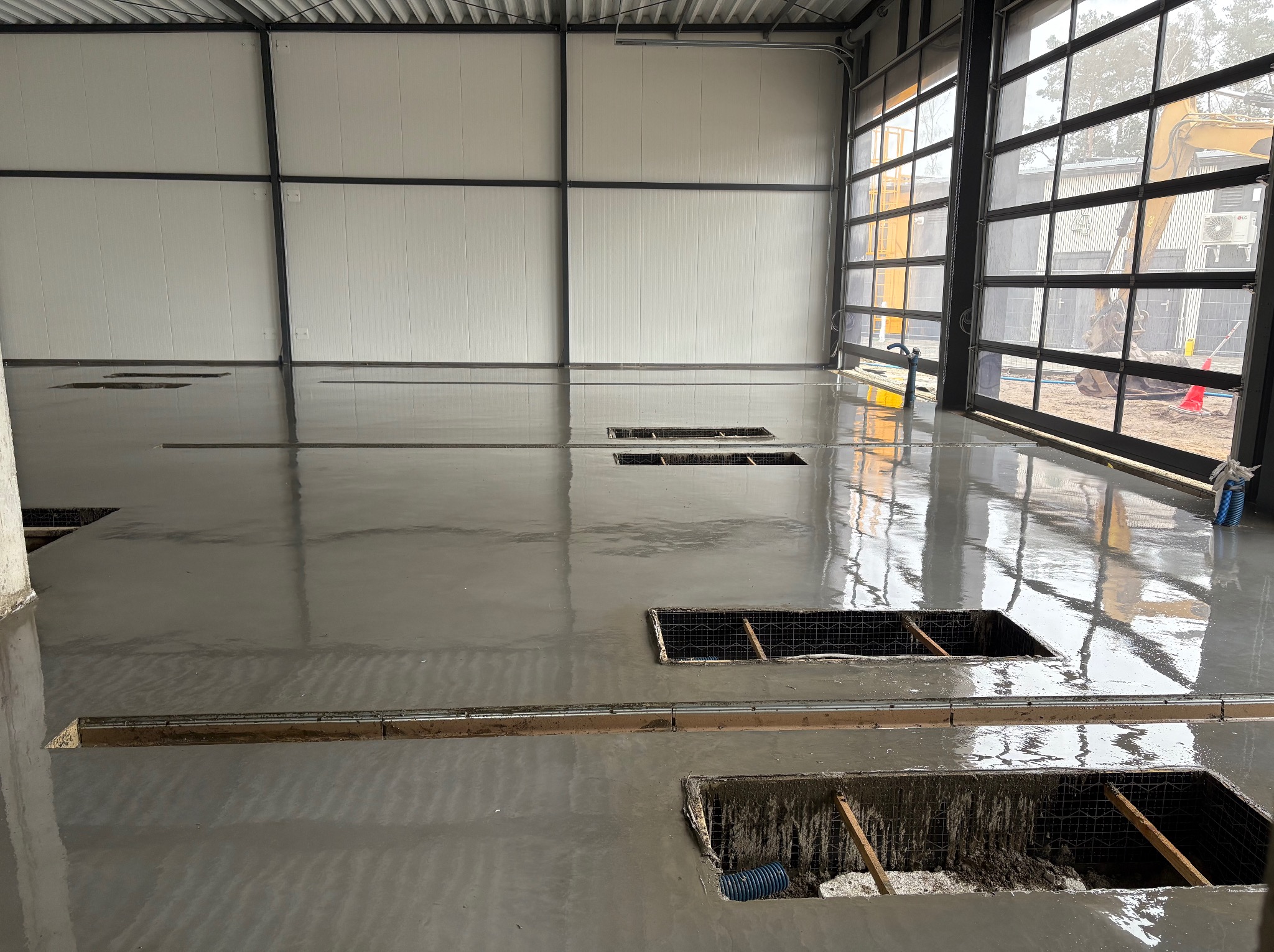 Świeżo wylana, błyszcząca posadzka w hali przemysłowej z widocznymi kratkami ściekowymi i otwartymi kanałami, z maszyną budowlaną widoczną przez otwarte drzwi.