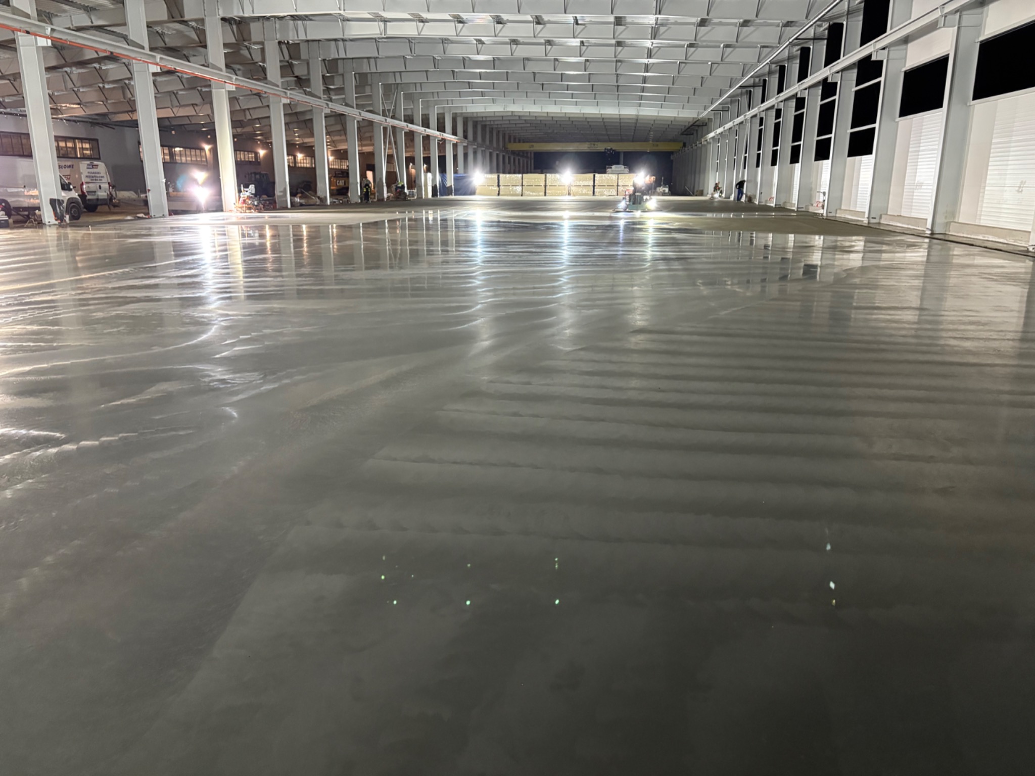 Wnętrze hali z nową, mokrą betonową posadzką w trakcie prac wykończeniowych; widoczne maszyny i ekipa budowlana w oddali.