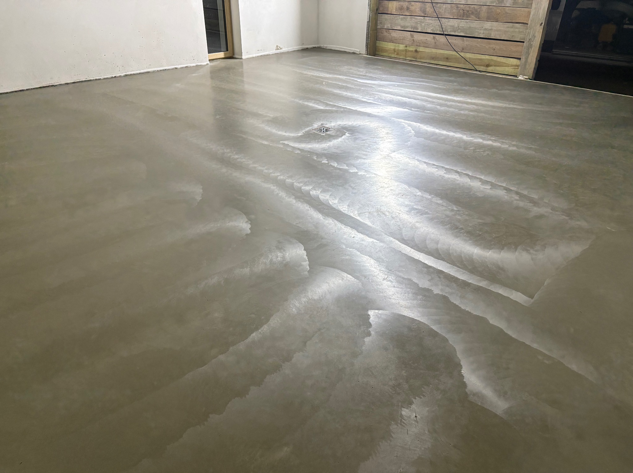 Świeżo wylana, gładka, szara posadzka z odpływem liniowym w pomieszczeniu w Szczutowie. Widoczne ślady po rozprowadzaniu masy, surowe wykończenie ścian.