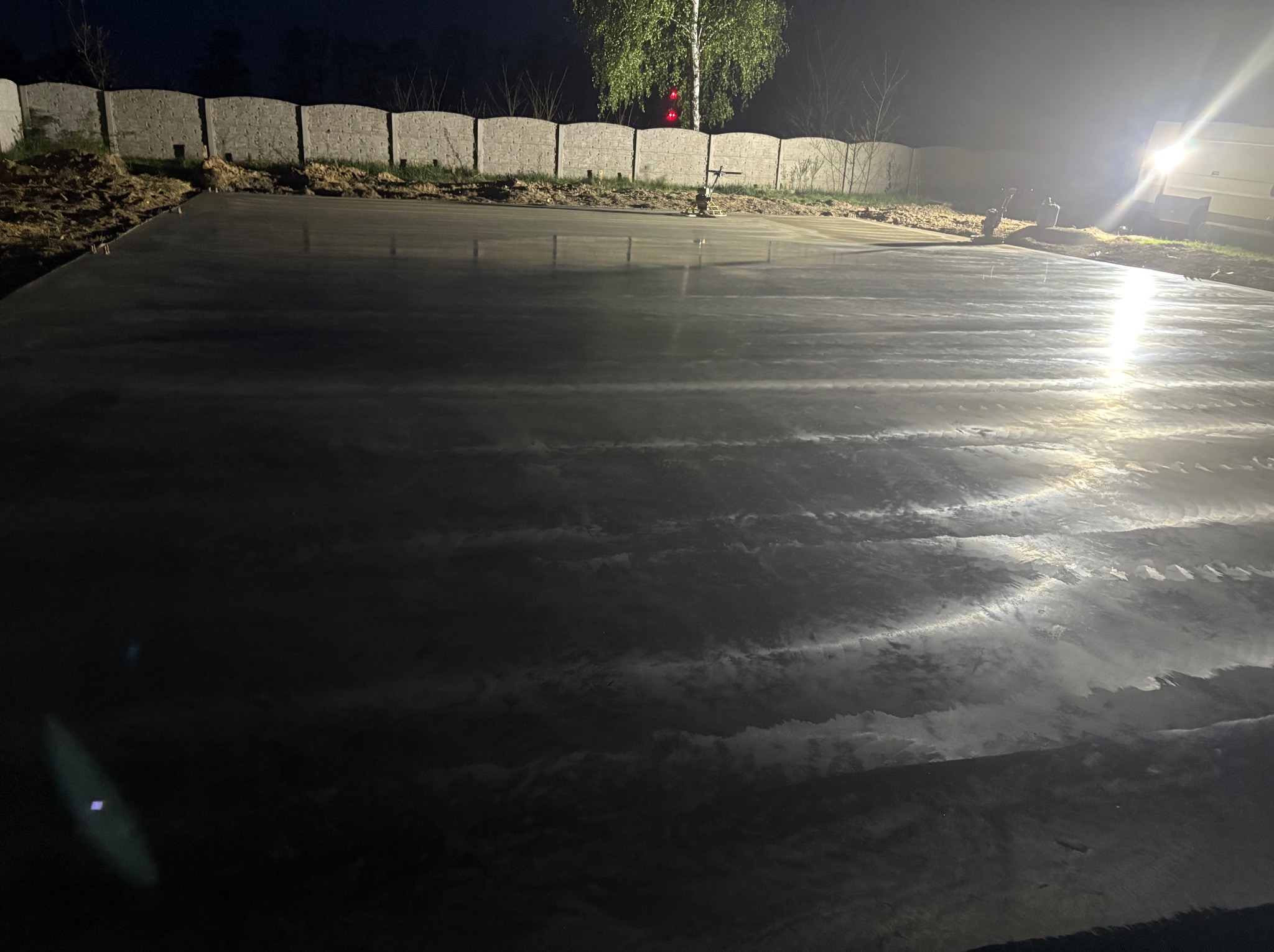 Świeżo wylana, mokra posadzka przemysłowa oświetlona nocą, zacieraczka obok. Widoczne ogrodzenie z prefabrykatów betonowych i drzewo. Nierównomierne odbicia światła.