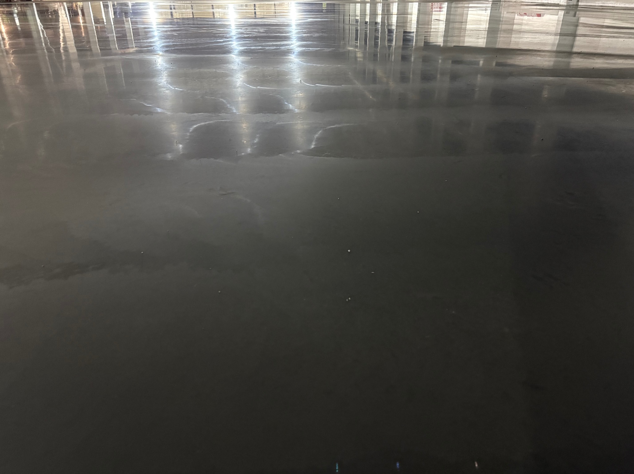 Błyszcząca, gładka, szara posadzka przemysłowa z refleksami świetlnymi. Widoczne kolumny konstrukcyjne w tle. Nowoczesna, minimalistyczna powierzchnia.