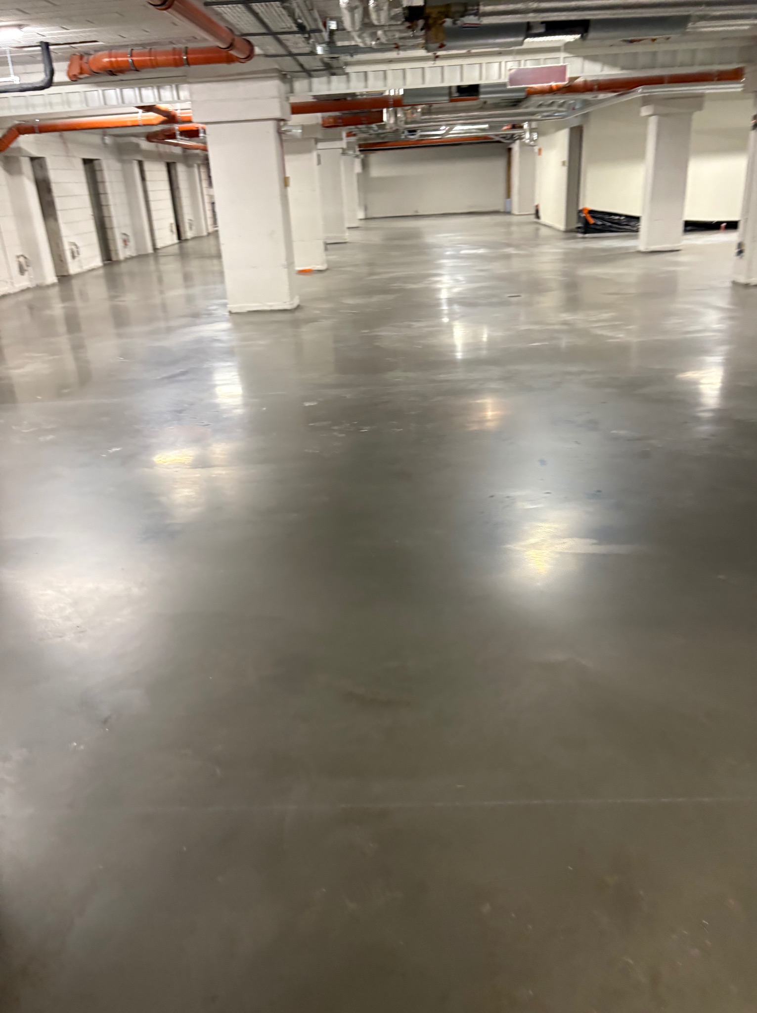 Błyszcząca, szara posadzka przemysłowa w hali z białymi kolumnami i pomarańczowymi rurami pod sufitem. Widoczne refleksy świetlne na gładkiej powierzchni.