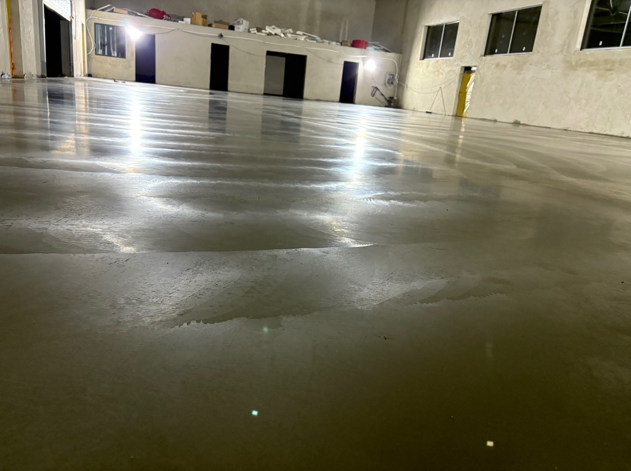 Wnętrze hali z nową, błyszczącą posadzką przemysłową w odcieniach szarości. Widoczne refleksy światła na gładkiej powierzchni i fragmenty ścian z oknami.
