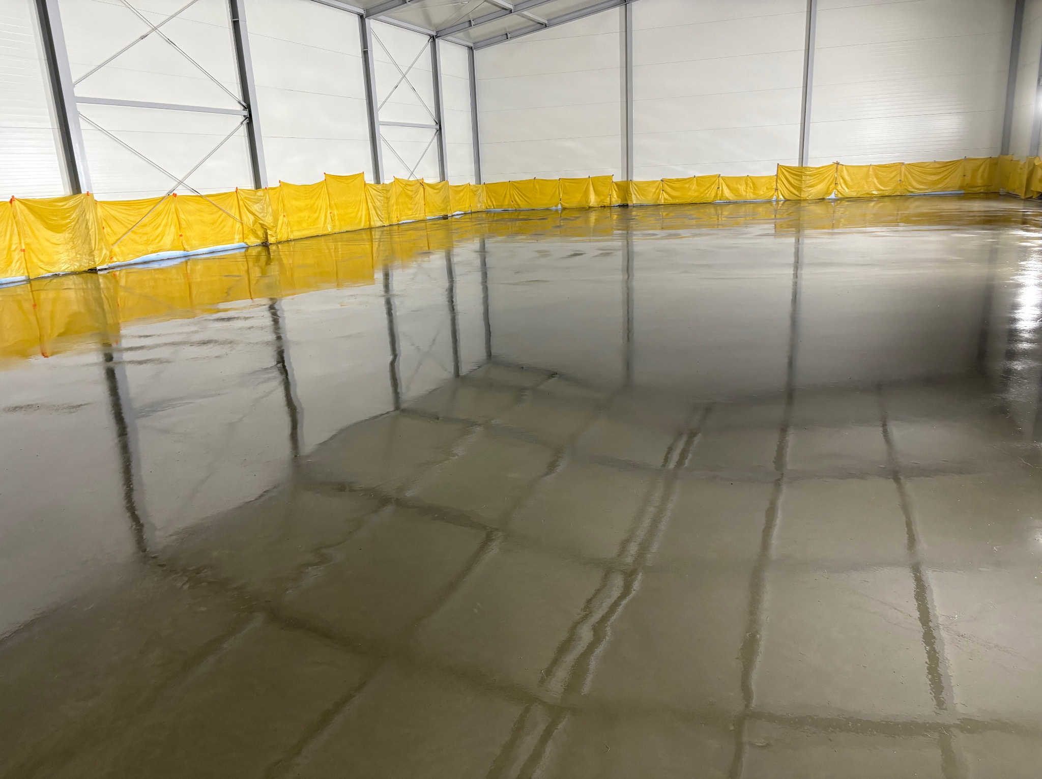 Świeżo wylana, błyszcząca posadzka w hali przemysłowej, zabezpieczona żółtymi osłonami przy ścianach. Widoczne refleksy światła na gładkiej powierzchni.